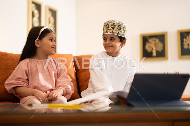 مراجعة الدروس اليومية عبر الانترنت، تطور العلم والتعليم في سلطنه عمان، بورتريه لتلميذين عربيين خليجيين عمانييين يرتدون الزي العماني التقليدي ويجلسون في غرفه المعيشه امام جهاز التابلت المحمول يقومون بحل الواجبات المدرسيه وتلقي المحاضرات المدرسيه عن بعد