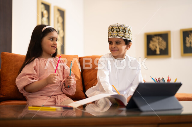 مراجعة الدروس اليومية عبر الانترنت، تطور العلم والتعليم في سلطنه عمان، بورتريه لتلميذين عربيين خليجيين عمانييين يرتدون الزي العماني التقليدي ويجلسون في غرفه المعيشه امام جهاز التابلت المحمول يقومون بحل الواجبات المدرسيه وتلقي المحاضرات المدرسيه عن بعد