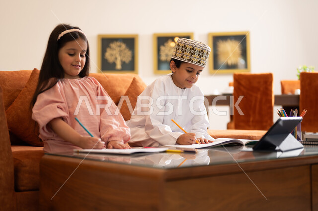 مراجعة الدروس اليومية عبر الانترنت، تطور العلم والتعليم في سلطنه عمان، بورتريه لتلميذين عربيين خليجيين عمانييين يرتدون الزي العماني التقليدي ويجلسون في غرفه المعيشه امام جهاز التابلت المحمول يقومون بحل الواجبات المدرسيه وتلقي المحاضرات المدرسيه عن بعد