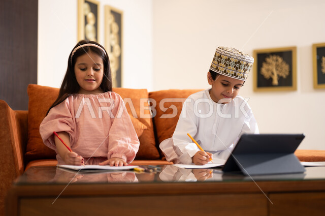 مراجعة الدروس اليومية عبر الانترنت، تطور العلم والتعليم في سلطنه عمان، بورتريه لتلميذين عربيين خليجيين عمانييين يرتدون الزي العماني التقليدي ويجلسون في غرفه المعيشه امام جهاز التابلت المحمول يقومون بحل الواجبات المدرسيه وتلقي المحاضرات المدرسيه عن بعد