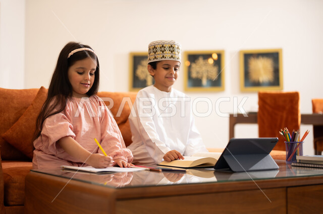 مراجعة الدروس اليومية عبر الانترنت، تطور العلم والتعليم في سلطنه عمان، بورتريه لتلميذين عربيين خليجيين عمانييين يرتدون الزي العماني التقليدي ويجلسون في غرفه المعيشه امام جهاز التابلت المحمول يقومون بحل الواجبات المدرسيه وتلقي المحاضرات المدرسيه عن بعد