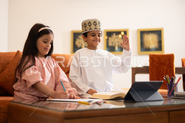 مراجعة الدروس اليومية عبر الانترنت، تطور العلم والتعليم في سلطنه عمان، بورتريه لتلميذين عربيين خليجيين عمانييين يرتدون الزي العماني التقليدي ويجلسون في غرفه المعيشه امام جهاز التابلت المحمول يقومون بحل الواجبات المدرسيه وتلقي المحاضرات المدرسيه عن بعد