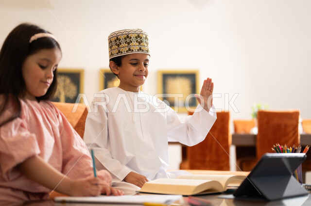 مراجعة الدروس اليومية عبر الانترنت، تطور العلم والتعليم في سلطنه عمان، بورتريه لتلميذين عربيين خليجيين عمانييين يرتدون الزي العماني التقليدي ويجلسون في غرفه المعيشه امام جهاز التابلت المحمول يقومون بحل الواجبات المدرسيه وتلقي المحاضرات المدرسيه عن بعد