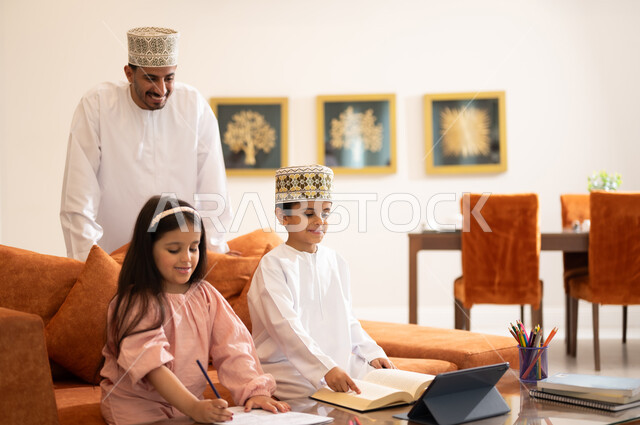 مفهوم التعليم المنزلي، مراجعة الدروس اليومية من خلال الاجهزة الالكترونية، بورتريه لطفل وطفله عربيين خليجيين عمانيان يرتديان الزي العماني التقليدي يجلسان في غرفه المعيشه أمام الجهاز اللوحي التعليمي ويقوم الاب بالاشراف عليهما ومساعدتهما في حل الواجبات المدرسيه