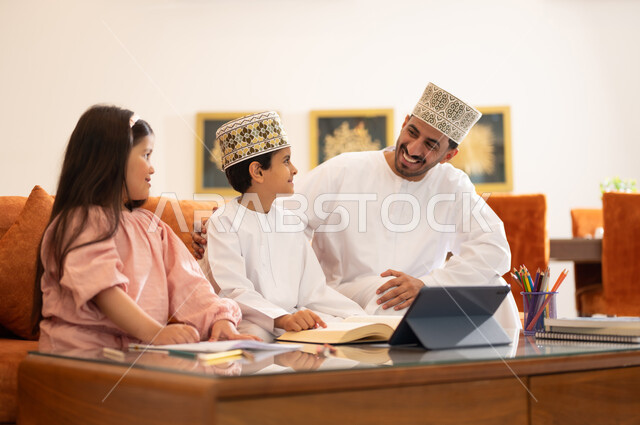 مفهوم التعليم المنزلي، مراجعة الدروس اليومية من خلال الاجهزة الالكترونية، بورتريه لطفل وطفله عربيين خليجيين عمانيان يرتديان الزي العماني التقليدي يجلسان بجوار والدهما في غرفه المعيشه أمام الجهاز اللوحي التعليمي، المذاكره والدراسه عن بعد