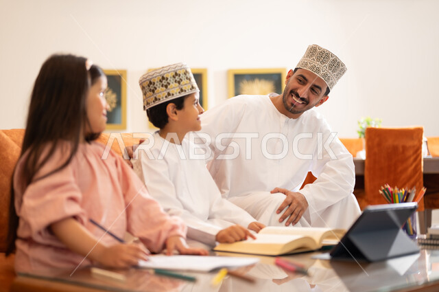 مفهوم التعليم المنزلي، مراجعة الدروس اليومية من خلال الاجهزة الالكترونية، بورتريه لطفل وطفله عربيين خليجيين عمانيان يرتديان الزي العماني التقليدي يجلسان بجوار والدهما في غرفه المعيشه أمام الجهاز اللوحي التعليمي، المذاكره والدراسه عن بعد