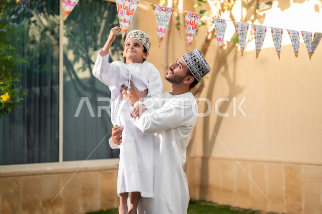 إظهار مشاعر الفرح والبهجة، حنان الأبوة ومشاعر المحبة والمودة، بورتريه لعائلة عربية خليجية عمانية تحتفل بالأعياد الإسلامية, أب يحمل ولده الصغير ليقوم بتعليق حبال الزينة داخل الحي السكني الخاص بهما، طقوس الاحتفالات والمناسبات السعيدة، ارتداء ملابس جديدة