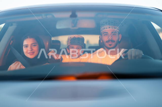 رحله جماعيه عائليه في السياره، دردشه ومحادثات في الطريق، التنزه في شوارع سلطنه عمان بورتريه لعائله عربيه خليجيه عمانيه مكونه من رجل وامرأه وصبي يرتديان الزي العماني التقليدي يجلسون داخل السياره ويلبسون احزمه الامان يقومون بجوله ترفيهيه، ايماءات تدل على السعاده والسرور وقضاء أوقات ممتعه