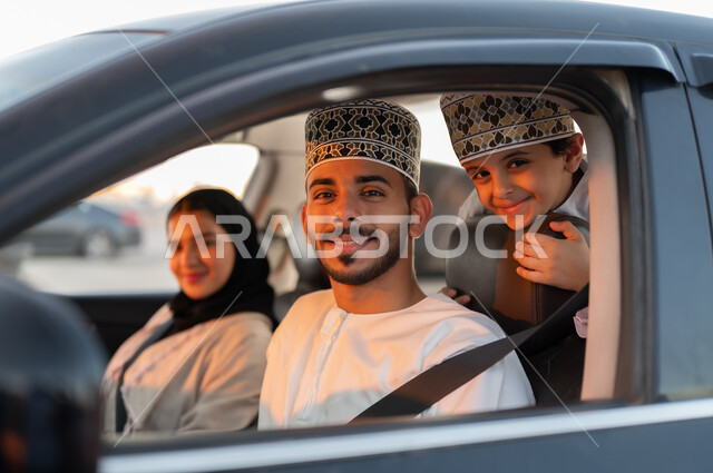 رحله جماعيه عائليه في السياره، دردشه ومحادثات في الطريق، التنزه في شوارع سلطنه عمان بورتريه لعائله عربيه خليجيه عمانيه مكونه من رجل وامرأه وصبي يرتديان الزي العماني التقليدي يجلسون داخل السياره ويلبسون احزمه الامان يقومون بجوله ترفيهيه، ايماءات تدل على السعاده والسرور وقضاء أوقات ممتعه