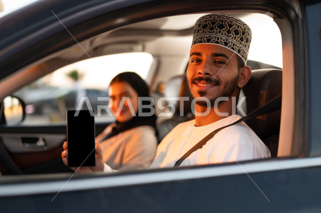 رحله جماعيه عائليه في السياره، دردشه ومحادثات في الطريق، قضاء وقت ممتع مع العائله، بورتريه لرجل عماني يرتدي الدشداشه والكمه يجلس بجوار زوجته في مقاعد السياره الاماميه يشير بهاتفهه المحمول بشاشه سوداء فارغه، القياده بحذر اثناء الانشغال بغيرالطريق