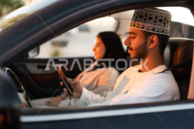 التنزه في شوارع سلطنه عمان، رحله جماعيه عائليه في السياره، دردشه ومحادثات في الطريق، قضاء وقت ممتع مع العائله، بورتريه لرجل عماني يرتدي الدشداشه والكمه يجلس بجوار زوجته في مقاعد السياره الاماميه ممسكا بيده هاتفهه المحمول، القياده بحذر اثناء الانشغال بغيرالطريق