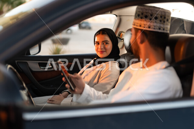 Walking in the streets of the Sultanate of Oman, a group family trip in the car, chatting and conversations on the way, spending quality time with the family, a portrait of an Omani man wearing a dishdasha and a cuff talking to his wife sitting next to him, holding his mobile phone in his hand, driving with caution while busy off the road.