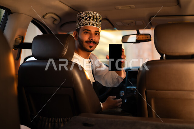The concept of driving in the Sultanate of Oman, riding in the car and looking at the back, a portrait of a smiling Omani Gulf Arab man wearing a dishdasha and a cuff sitting on the driving seat holding his mobile phone and pointing at it with a blank black screen, using mobile applications and GPS to access websites easily and smoothly.