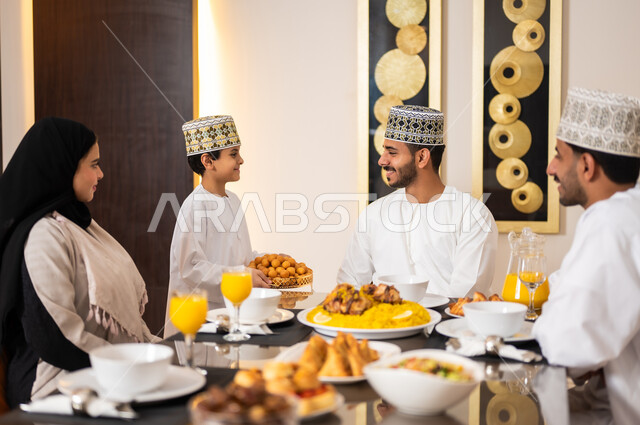 تجهيز وتحضير سفرة الإفطار، اجتماع الأسرة حول طاولة الطعام المليئه بأصناف المأكولات و المقبلات الشهية، ديكورات وأجواء رمضانية، بورتريه لعائله عربيه خليجيه عمانيه ترتدي الزي العماني التقليدي مجتمعين حول مائده الطعام في انتظار مدفع الافطار