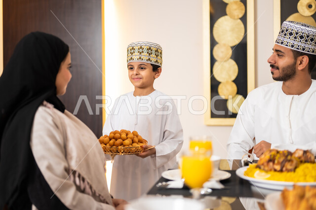 Preparing the Iftar meal, the family gathering around the dining table filled with delicious food and appetizers, Ramadan decorations and atmosphere, a portrait of an Arab Gulf Omani family wearing traditional Omani dress, gathered around the dining table sharing breakfast with each other.