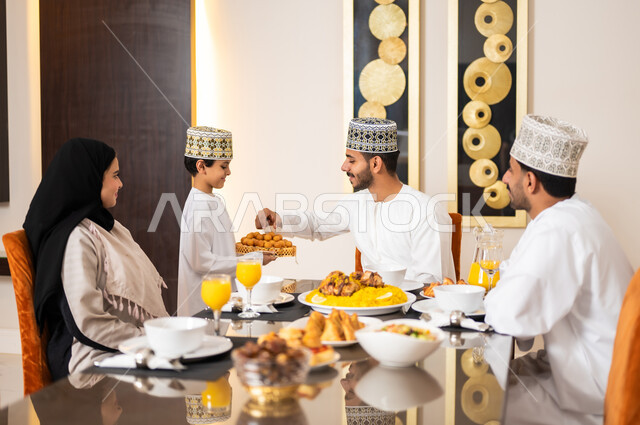 Preparing and preparing the breakfast table, the family gathering around the dining table filled with delicious food and appetizers, Ramadan decorations and atmosphere, a portrait of an Arab Gulf Omani family wearing traditional Omani dress, gathered around the dining table waiting for the breakfast cannon.
