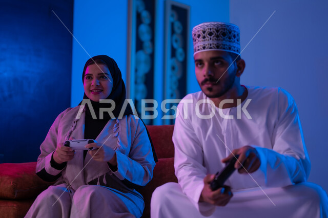 مفهوم التسليه والترفيه الجماعي مع الاهل, بورتريه لرجل وأمراه عربين خليجين عمانيين يرتديان الزي العماني التقليدي يجلسان على أريكه مريحه في غرفه المعيشه يحملان بيدهما عصا التحكم بالالعاب الالكترونيه يتحديان بعضهما للفوز بالجوله, تعابير تدل على الحماس والسرور، الحزن والخساره