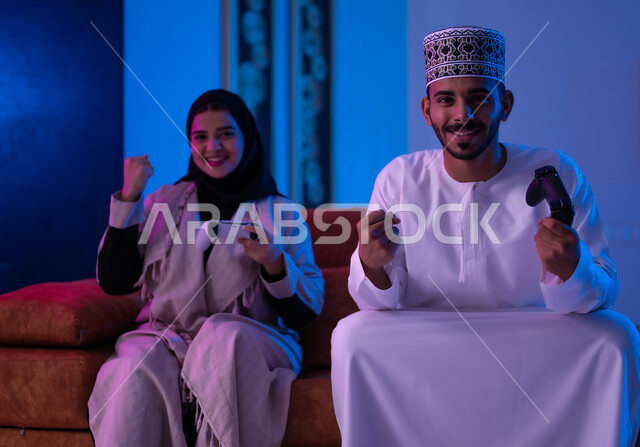 The concept of group entertainment and recreation with family, a portrait of an Omani Gulf Arab man and woman wearing traditional Omani clothing, sitting on a comfortable sofa in the living room, holding an electronic game control stick in their hands, challenging each other to win the round, expressions indicating enthusiasm and pleasure, sadness and loss.