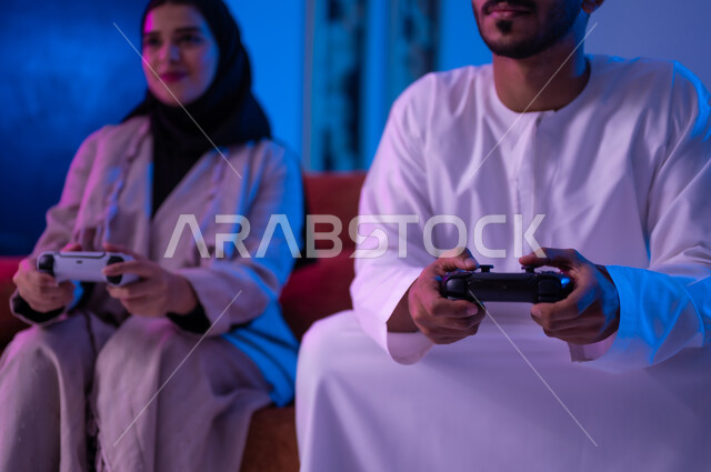 مفهوم التسليه والترفيه الجماعي مع الاهل, بورتريه لرجل وأمراه عربين خليجين عمانيين يرتديان الزي العماني التقليدي يجلسان على أريكه مريحه في غرفه المعيشه يحملان بيدهما عصا التحكم بالالعاب الالكترونيه يتحديان بعضهما للفوز بالجوله, تعابير تدل على الحماس والسرور، الحزن والخساره