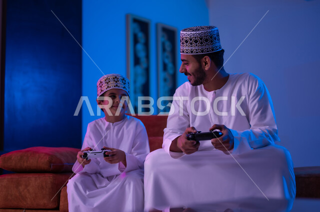 مفهوم التسليه والترفيه الجماعي مع الابناء, بورتريه لرجل وطفل عربين خليجين عمانيين يرتديان الدشداشة و الكمه يجلسان على أريكه مريحه في غرفه المعيشه يحملان بيدهما عصا التحكم بالالعاب الالكترونيه يتحديان بعضهما للفوز بالجوله, تعابير تدل على الحماس والسرور، الحزن والخساره