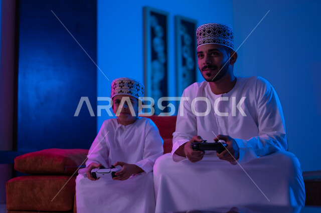 مفهوم التسليه والترفيه الجماعي مع الابناء, بورتريه لرجل وطفل عربين خليجين عمانيين يرتديان الدشداشة و الكمه يجلسان على أريكه مريحه في غرفه المعيشه يحملان بيدهما عصا التحكم بالالعاب الالكترونيه يتحديان بعضهما للفوز بالجوله, تعابير تدل على الحماس والسرور، الحزن والخساره