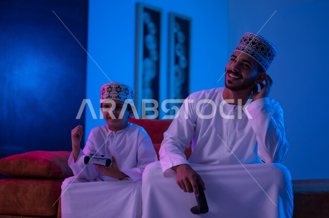 مفهوم التسليه والترفيه الجماعي مع الابناء, بورتريه لرجل وطفل عربين خليجين عمانيين يرتديان الدشداشة و الكمه يجلسان على أريكه مريحه في غرفه المعيشه يحملان بيدهما عصا التحكم بالالعاب الالكترونيه يتحديان بعضهما للفوز بالجوله, تعابير تدل على الحماس والسرور، الحزن والخساره