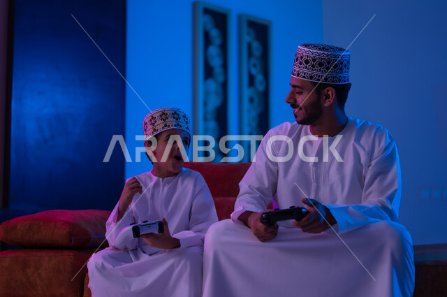 مفهوم التسليه والترفيه الجماعي مع الابناء, بورتريه لرجل وطفل عربين خليجين عمانيين يرتديان الدشداشة و الكمه يجلسان على أريكه مريحه في غرفه المعيشه يحملان بيدهما عصا التحكم بالالعاب الالكترونيه يتحديان بعضهما للفوز بالجوله, تعابير تدل على الحماس والسرور، الحزن والخساره