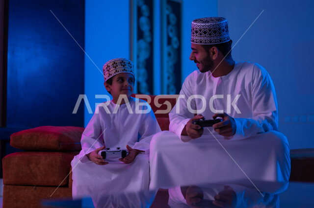 مفهوم التسليه والترفيه الجماعي مع الابناء, بورتريه لرجل وطفل عربين خليجين عمانيين يرتديان الدشداشة و الكمه يجلسان على أريكه مريحه في غرفه المعيشه يحملان بيدهما عصا التحكم بالالعاب الالكترونيه يتحديان بعضهما للفوز بالجوله, تعابير تدل على الحماس والسرور، الحزن والخساره