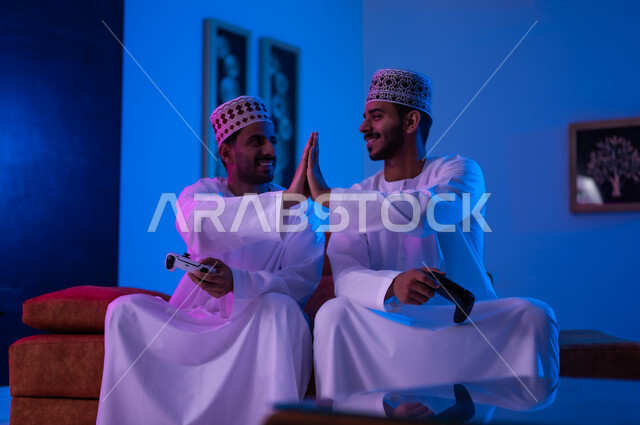 The concept of group entertainment and entertainment with friends, a portrait of two young Arab Gulf Omani men wearing a dishdasha and a kabbah, sitting on a comfortable sofa in the living room, holding an electronic game control stick in their hands, challenging each other to win the round, expressions indicating enthusiasm and pleasure, sadness and loss.