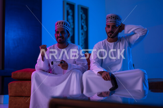 مفهوم التسليه والترفيه الجماعي مع الاصدقاء, بورتريه لشابين عربين خليجين عمانيين يرتديان الدشداشة و الكمه يجلسان على أريكه مريحه في غرفه المعيشه يحملان بيدهما عصا التحكم بالالعاب الالكترونيه يتحديان بعضهما للفوز بالجوله, تعابير تدل على الحماس والسرور، الحزن والخساره
