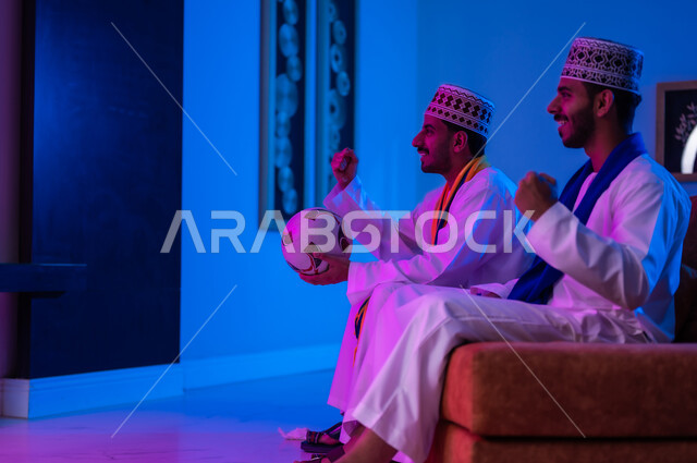 قضاء أوقات ممتعه بمشاهده مباريات كره القدم ، بورتريه لصديقين عربين خليجين عمانيين يرتديان الدشداشة و الكمه يجلسان على أريكه مريحه في غرفه المعيشه يقومان بمشاهده التلفاز، تعابيروجه ويدين تدلان على الحماس  والتشجيع، الشغف بمتابعه مباريات كره القدم وتمني الفوزللفريق