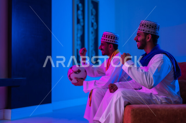 Spending enjoyable times watching football matches, a portrait of two Arab Gulf Omani friends wearing a dishdasha and a cuff, sitting on a comfortable sofa in the living room, watching television, their facial expressions and hands indicate enthusiasm and encouragement, the passion for watching football matches and wishing the team victory.