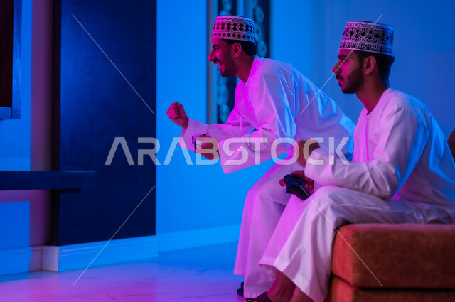 Spending enjoyable times watching football matches, a portrait of two Arab Gulf Omani friends wearing a dishdasha and a cuff, sitting on a comfortable sofa in the living room, watching television, their facial expressions and hands indicate enthusiasm and encouragement, the passion for watching football matches and wishing the team victory.