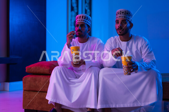 The concept of group entertainment and entertainment, spending a good time eating popcorn while watching television, a portrait of two Arab, Gulf, and Omani friends wearing a dishdasha and a kabbah, sitting on a comfortable sofa in the living room in front of the television screen, holding cups full of popcorn in their hands, expressions indicating happiness and contentment.