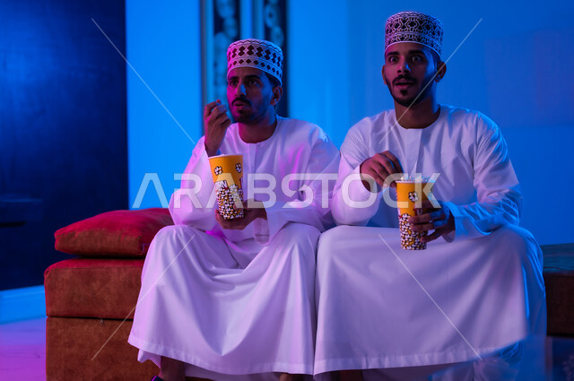 مفهوم التسليه والترفيه الجماعي, قضاء وقت ممتع بتناول الفوشار اثناء مشاهده التلفاز، بورتريه لصديقين عربين خليجين عمانيين يرتديان الدشداشة و الكمه يجلسان على أريكه مريحه في غرفه المعيشه امام شاشه التلفاز يحملان بيدهما أكواب مليئه بالبوب كورن، تعابير تدل على السعاده والسرور