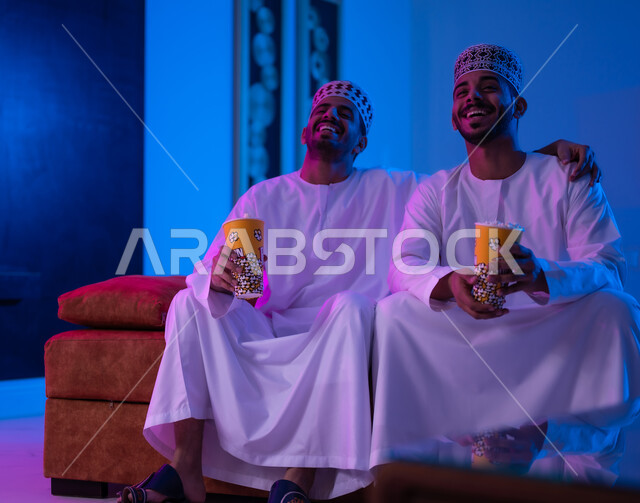 مفهوم التسليه والترفيه الجماعي, قضاء وقت ممتع بتناول الفوشار اثناء مشاهده التلفاز، بورتريه لصديقين عربين خليجين عمانيين يرتديان الدشداشة و الكمه يجلسان على أريكه مريحه في غرفه المعيشه امام شاشه التلفاز يحملان بيدهما أكواب مليئه بالبوب كورن، تعابير تدل على السعاده والسرور
