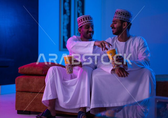 مفهوم التسليه والترفيه الجماعي, قضاء وقت ممتع بتناول الفوشار اثناء مشاهده التلفاز، بورتريه لصديقين عربين خليجين عمانيين يرتديان الدشداشة و الكمه يجلسان على أريكه مريحه في غرفه المعيشه امام شاشه التلفاز يحملان بيدهما أكواب مليئه بالبوب كورن، تعابير تدل على السعاده والسرور