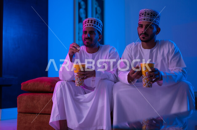 مفهوم التسليه والترفيه الجماعي, قضاء وقت ممتع بتناول الفوشار اثناء مشاهده التلفاز، بورتريه لصديقين عربين خليجين عمانيين يرتديان الدشداشة و الكمه يجلسان على أريكه مريحه في غرفه المعيشه امام شاشه التلفاز يحملان بيدهما أكواب مليئه بالبوب كورن، تعابير تدل على السعاده والسرور