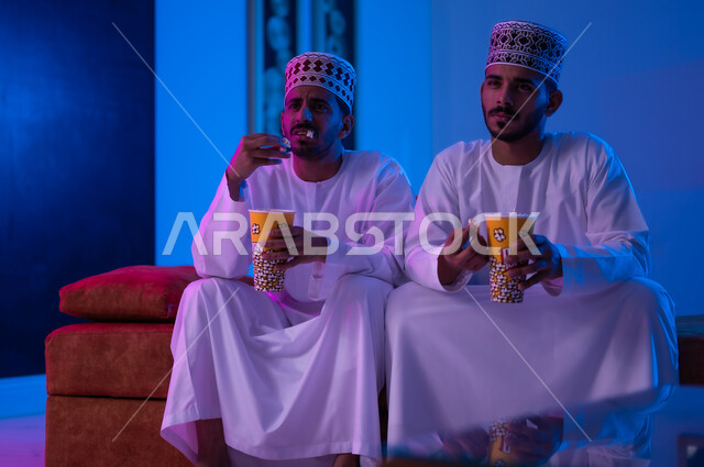 مفهوم التسليه والترفيه الجماعي, قضاء وقت ممتع بتناول الفوشار اثناء مشاهده التلفاز، بورتريه لصديقين عربين خليجين عمانيين يرتديان الدشداشة و الكمه يجلسان على أريكه مريحه في غرفه المعيشه امام شاشه التلفاز يحملان بيدهما أكواب مليئه بالبوب كورن، تعابير تدل على السعاده والسرور