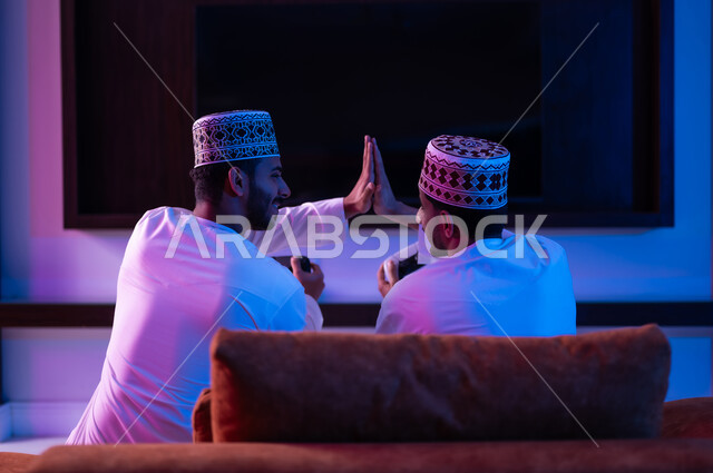 مفهوم التسليه والترفيه الجماعي, بورتريه لشابين عربين خليجين عمانيين يرتديان الدشداشة و الكمه يجلسان على أريكه مريحه في غرفه المعيشه امام شاشه التلفاز يحملان بيدهما عصا التحكم بالالعاب الالكترونيه يتحديان بعضهما للفوز بالجوله, تعابير تدل على الحماس والسرور
