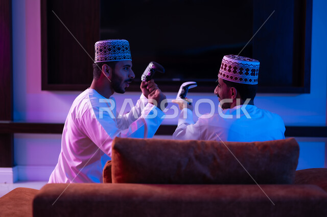 The concept of group entertainment and entertainment, a portrait of two young Arab Gulf Omani men wearing a dishdasha and a kabbah, sitting on a comfortable sofa in the living room in front of the TV screen, holding an electronic game control stick in their hands, challenging each other to win the round, expressions indicating enthusiasm and pleasure.