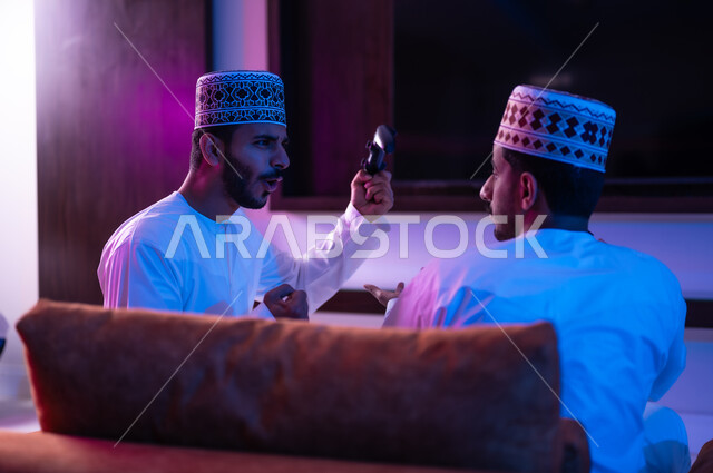 The concept of group entertainment and entertainment, a portrait of two young Arab Gulf Omani men wearing a dishdasha and a kabbah, sitting on a comfortable sofa in the living room in front of the TV screen, holding an electronic game control stick in their hands, challenging each other to win the round, expressions indicating enthusiasm and pleasure.