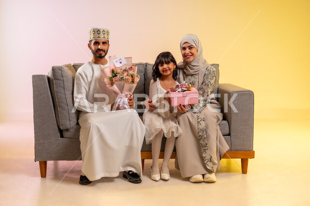 الاحتفال بقدوم العيد بالهدايا والأزهار، تقدير الوالدين والبر بهم، المودة والمحبة بين أفراد الأسرة، تقوية الروابط الزوجية، بورتريه لرجل عربي خليجي عماني يرتدي الدشداشة والكمه يقوم بتقديم الورود لزوجته برفقه ابنته، عائلة عمانية تحتفل بسعادة، بخلفية ملونة