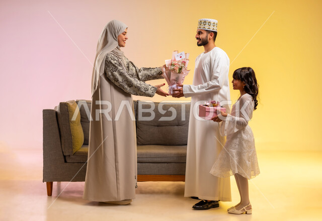 تقديم هدايا العيد للأم والمفاجآت، المودة والمحبة بين أفراد الأسرة، تقوية الروابط الزوجية، بورتريه لرجل عربي خليجي عماني يرتدي الدشداشة والكمه يقوم بتقديم الورود لزوجته برفقه ابنته، عائلة عمانية تقوم بالاحتفال بقدوم العيد بإيماءات السعادة، خلفية ملونة
