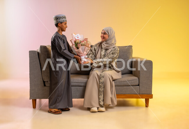 مفهوم بر الوالدين والإهداء، مشاعر الحب والحنان بمنح الأم هدايا العيد، أجواء لمة الأهل في المناسبات السعيدة, بورتريه لولد عربي خليجي عماني يرتدي الدشداشة والكمه يقوم بإعطاء أمه باقة من الورد بإيماءات السعادة, عائلة عمانية تحتفل بالعيد المبارك, خلفية ملونة