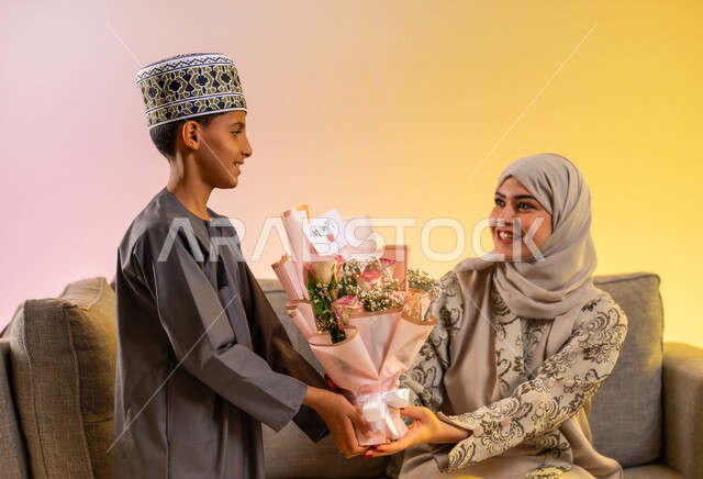 مشاعر الحب والحنان بمنح الأم هدايا العيد، تقدير الوالدين ومفهوم الإهداء, أجواء لمة الأهل في المناسبات السعيدة, بورتريه لولد عربي خليجي عماني يرتدي الدشداشة والكمه يقوم بإعطاء أمه باقة من الورد بإيماءات السعادة, عائلة عمانية تحتفل بالعيد المبارك, خلفية ملونة