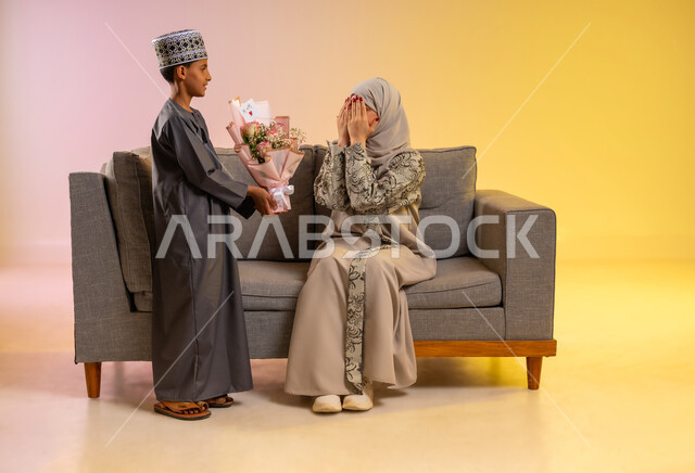 منح الأم هدايا العيد، تقدير الوالدين ومفهوم الإهداء, أجواء لمة الأهل في المناسبات السعيدة, بورتريه لولد عربي خليجي عماني يرتدي الدشداشة والكمه يقوم بإعطاء أمه باقة من الورد الملون بإيماءات السعادة, عائلة عمانية تحتفل بالعيد المبارك, خلفية ملونة