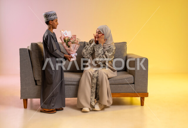 إيماءات المفاجأة بهدايا العيد, أجواء لمة الأهل في المناسبات السعيدة, بورتريه لولد عربي خليجي عماني يرتدي الدشداشة والكمه يقوم بإعطاء أمه باقة من الورد الملون, مفهوم الإهداء وتقدير الوالدين, عائلة عمانية تحتفل بالعيد المبارك, خلفية ملونة