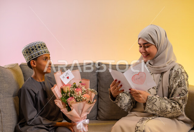 هدايا العيد وكروت المعايدة, بورتريه لولد عربي خليجي عماني يرتدي الدشداشة والكمه يقوم بإعطاء أمه باقة من الورد ورسالة مكتوبة يدوياً, مفهوم الإهداء وتقدير الوالدين, عائلة عمانية تحتفل بالعيد المبارك, أجواء لمة الأهل في المناسبات والاحتفالات السعيدة, خلفية ملونة