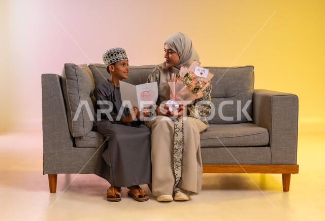كروت المعايدة وهدايا العيد, بورتريه لولد عربي خليجي عماني يرتدي الدشداشة والكمه يقوم بإعطاء أمه باقة من الورد الملون, مفهوم الإهداء وتقدير الوالدين, عائلة عمانية تحتفل بالعيد المبارك, أجواء لمة الأهل في المناسبات والاحتفالات السعيدة, خلفية ملونة